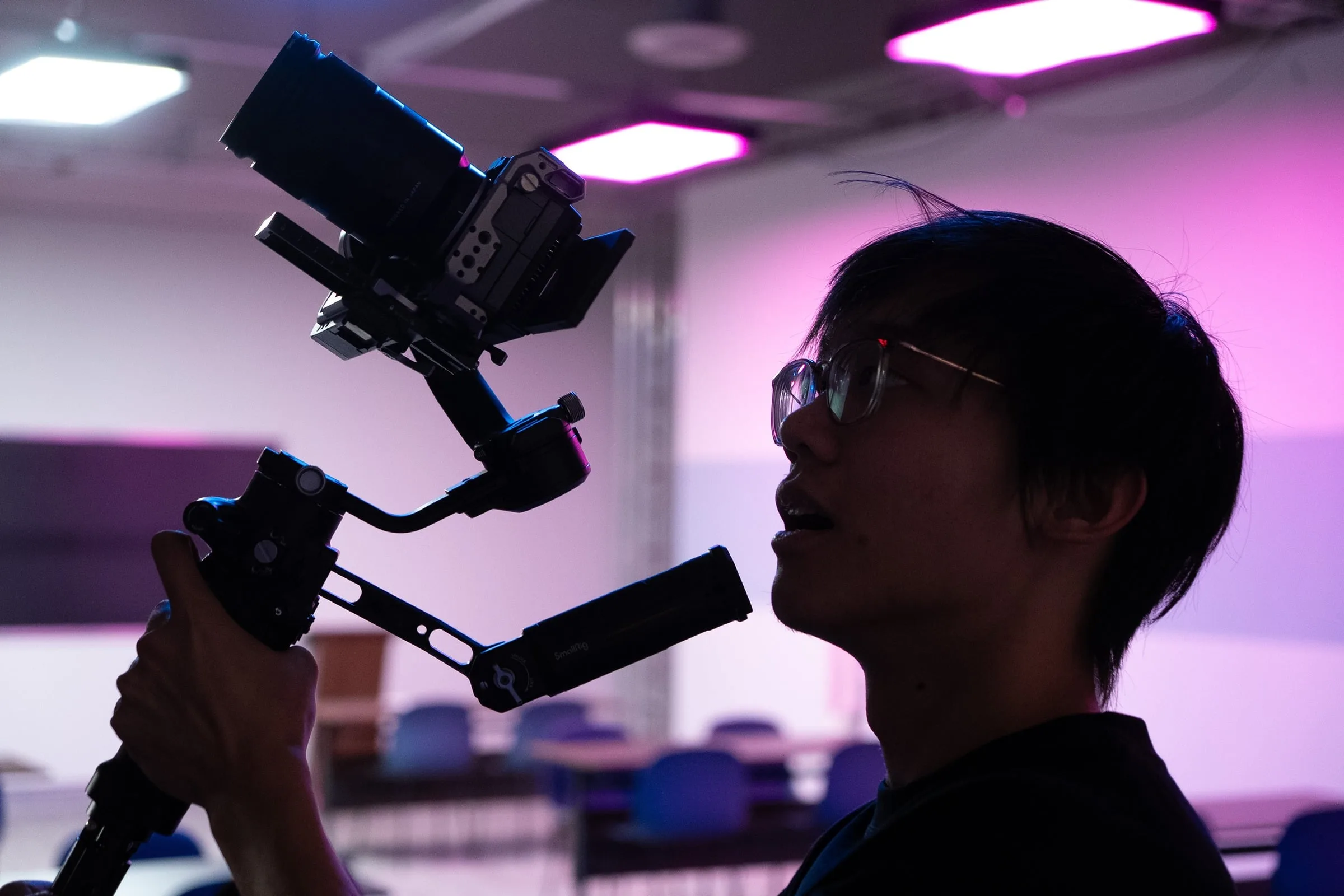 A camera operator holds a stabilized cinema camera under magenta studio lighting.