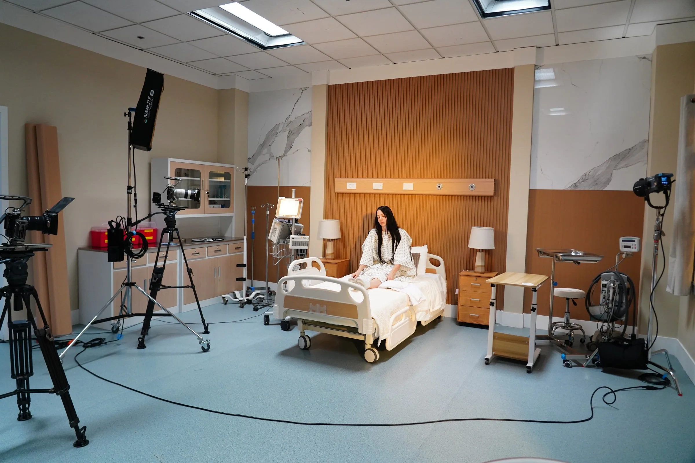 Cinema cameras and LED lights surround a performer on a practical hospital-room set.