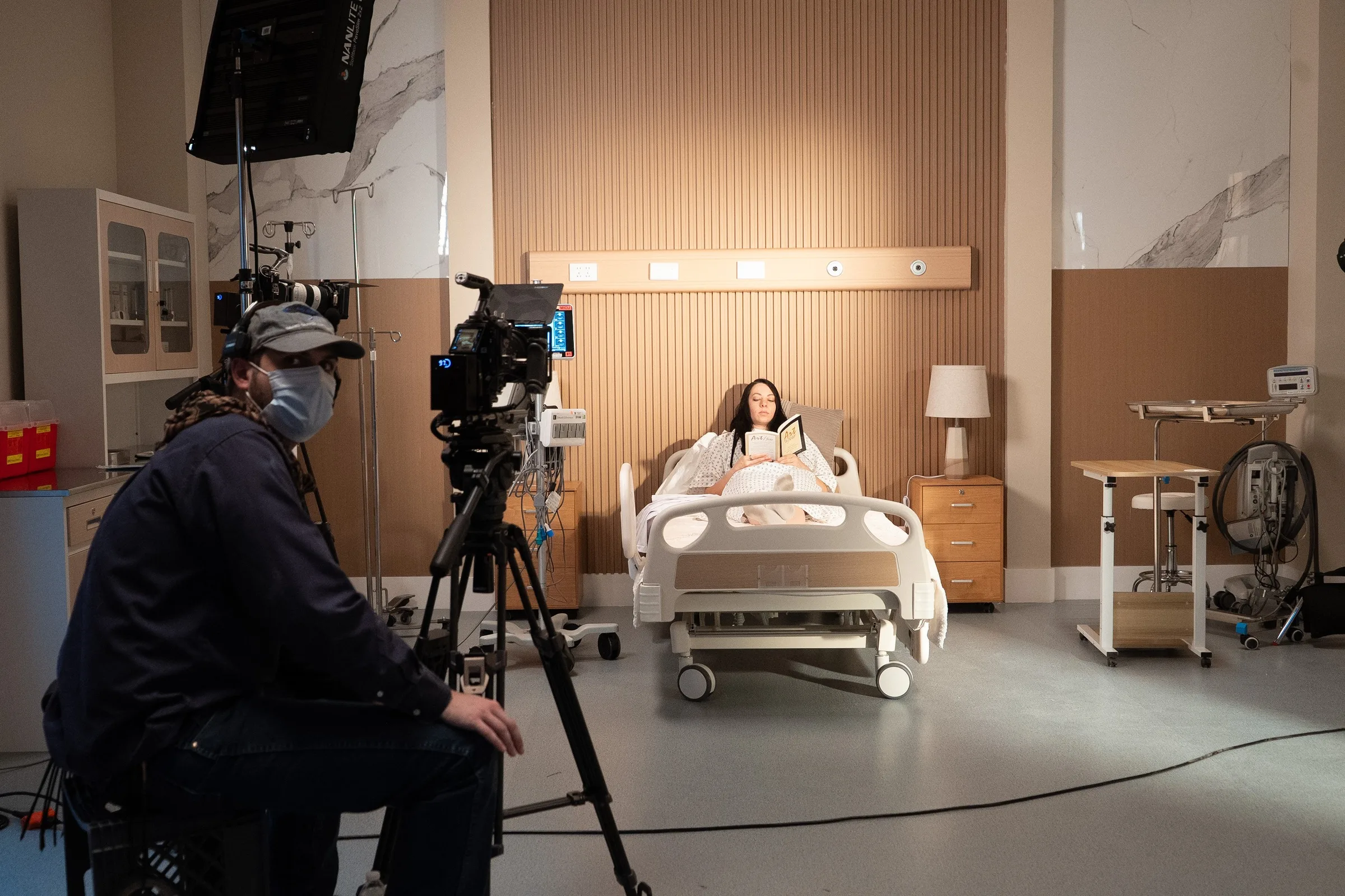 A camera operator frames a performer on a practical hospital-room set.