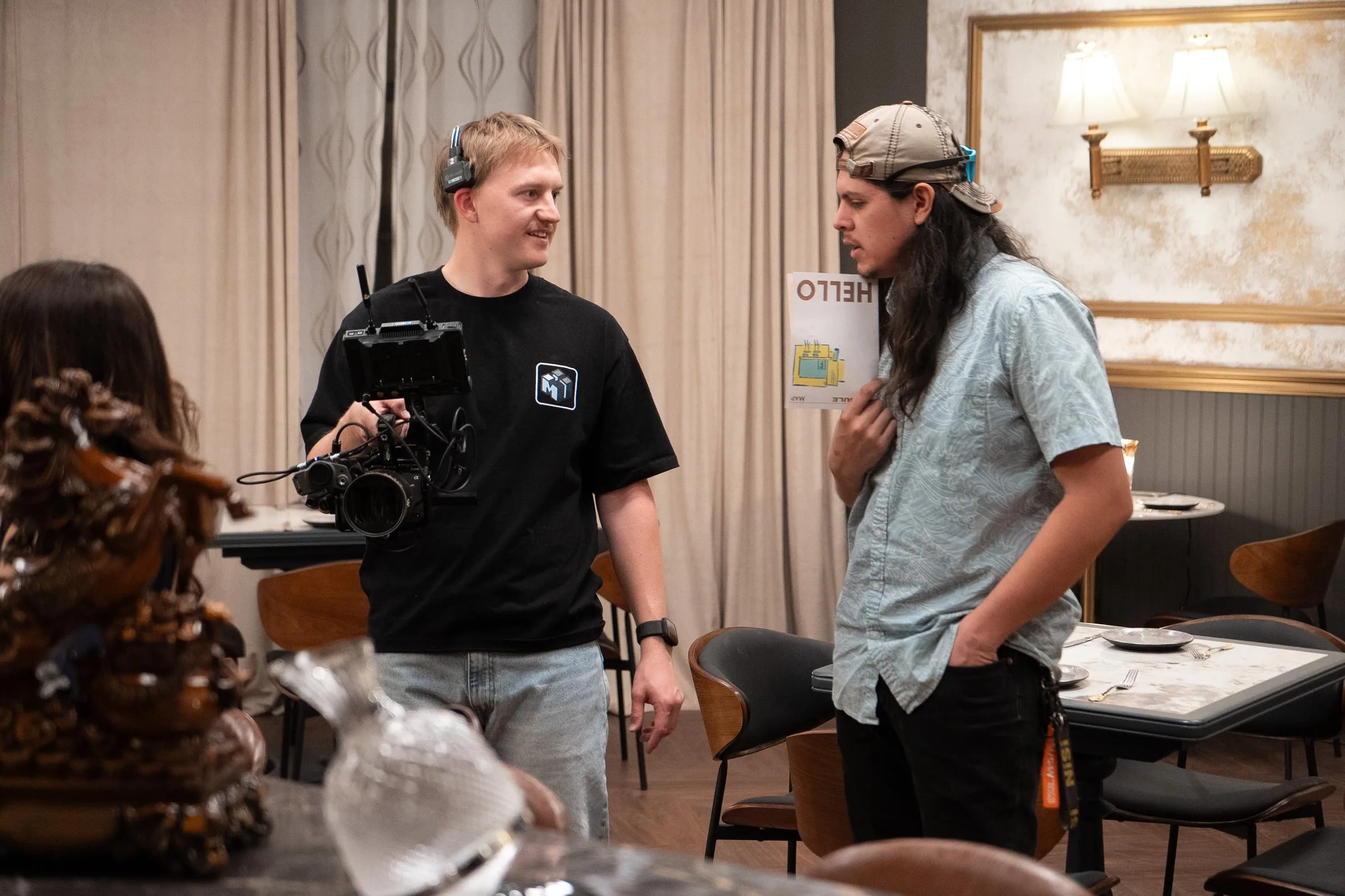 Two production crew members confer beside a cinema camera on a restaurant set.